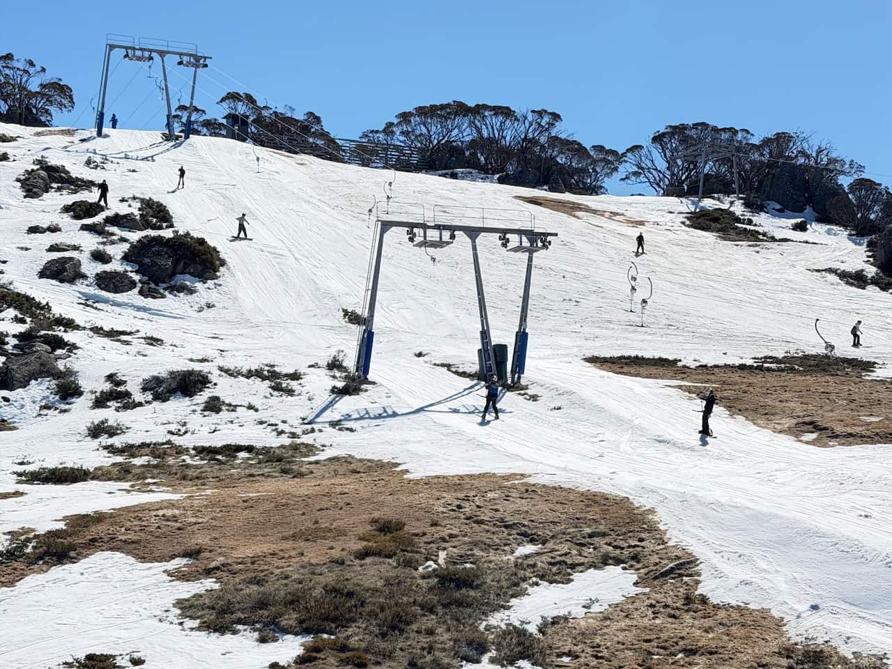 A ski slope from a distance with several people skiing and snowboarding. The snow is patchy with large sections of dirt and rock coming throw, a ski lift through the middle.