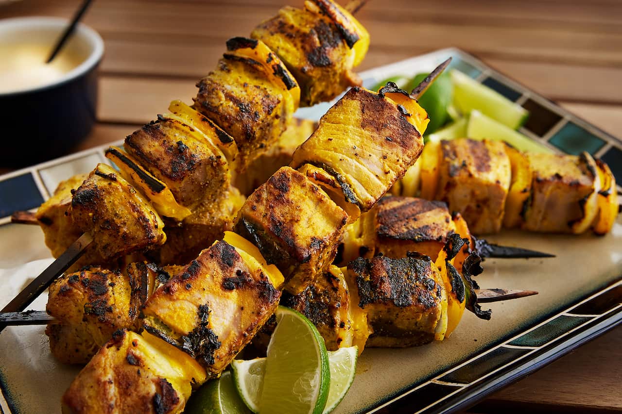 Five skewers, threaded with golden-coloured grilled tuna and onion, sit on a plate with lime wedges. 