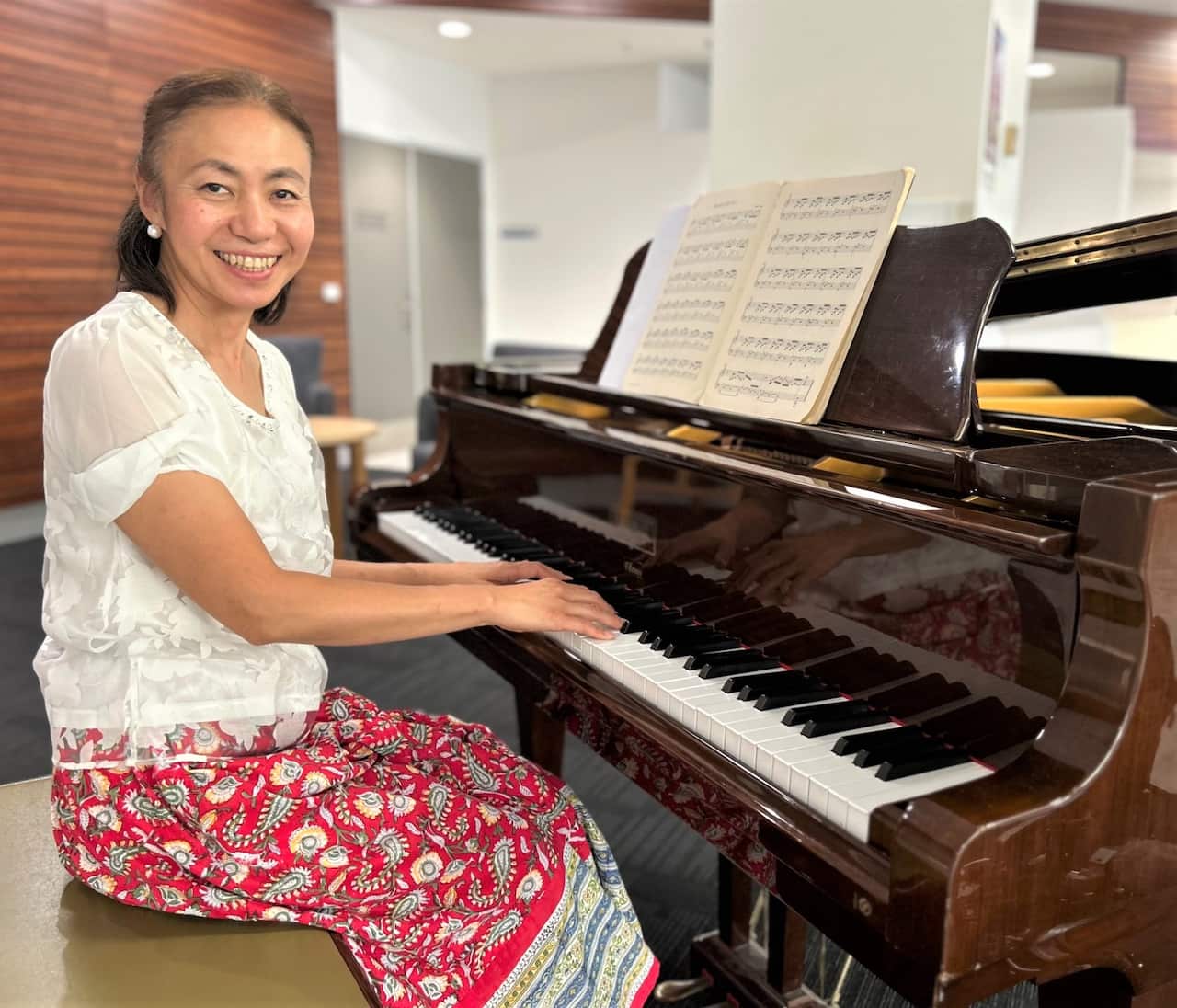 A woman in a white shirt and floral skirt sits at a piano keyboard smiling at camera.