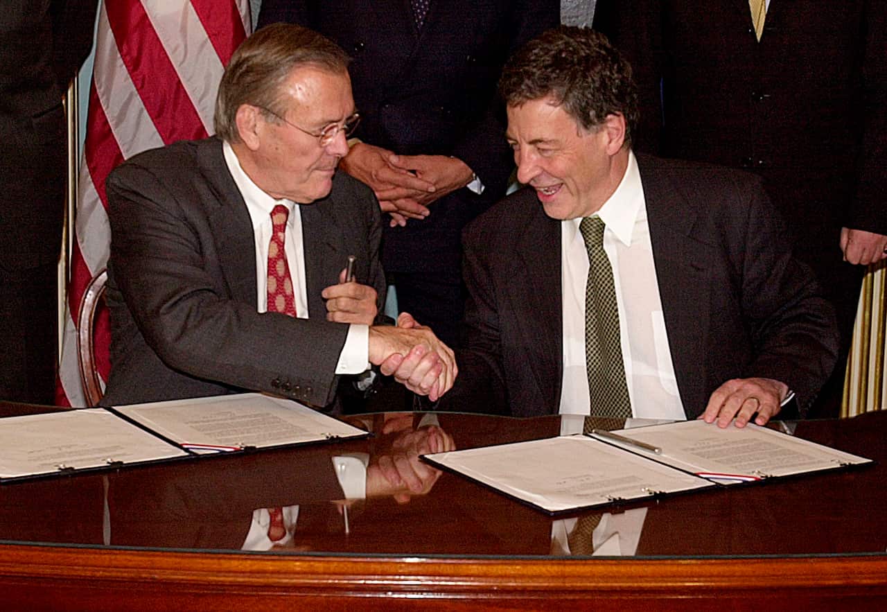 Two suited men shake hands at a table with documents in front of them.