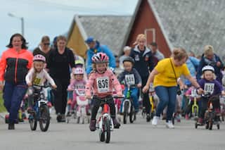 Children wearing helmets and bibs with numbers ride bikes while parents run alongside them.