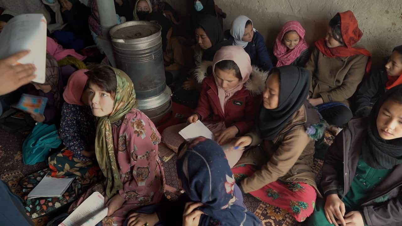 Teenage girls wearing headscarves sit on the floor in a crowded room holding open textbooks