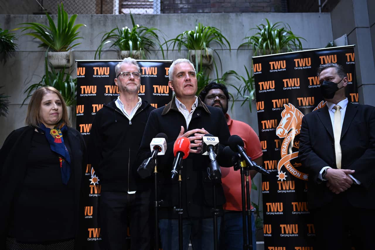 Five people standing in front of microphones and backdrops for a press conference.