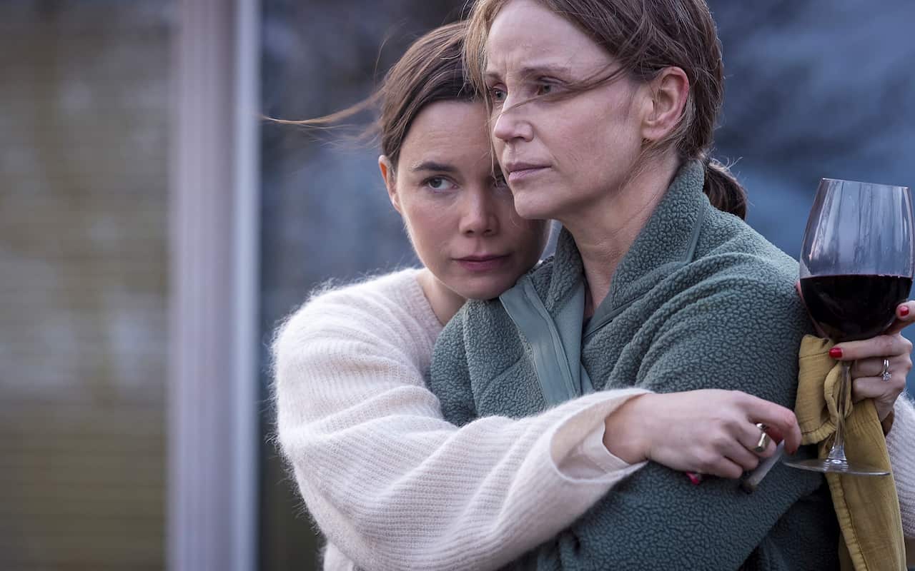 A woman in a white knit sweater, holding a glass of red wine in one hand, hugs another woman in a blueish fleece top. The gesture appears comforting, although the second woman is gazing off into the distance.