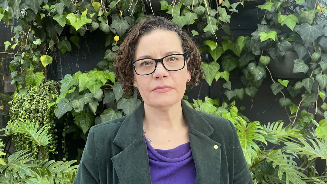 a woman with a curly bob and black glasses and wearing a green blazer stands in front of a green garden