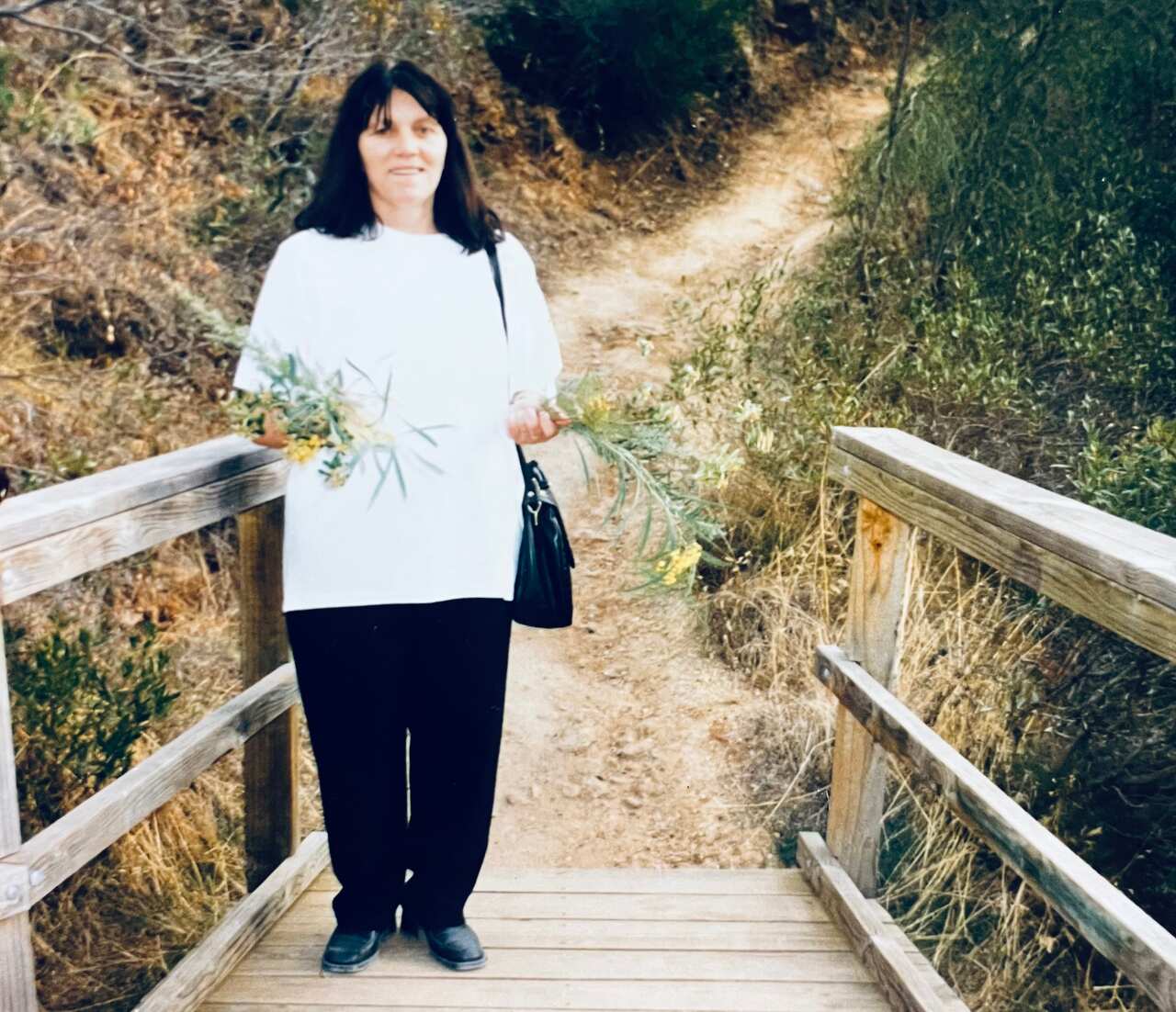 Jeka stands in a white t-shirt and black pants, holding a tuft of flowers on a wooden bridge. 