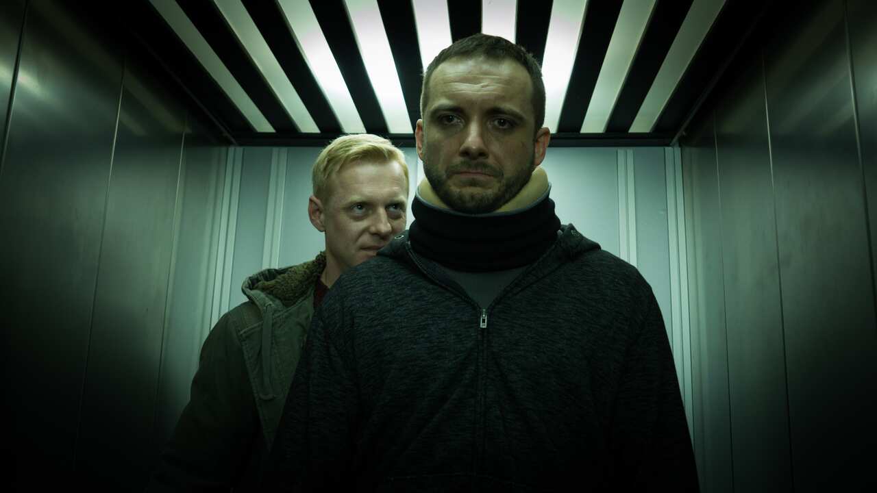 A man menacingly stands behind another man in an elevator