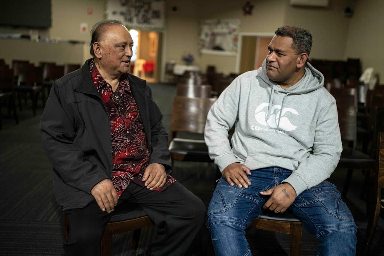A man in a red patterned shirt and black jacket sits beside another man wearing a grey hoodie and blue jeans.