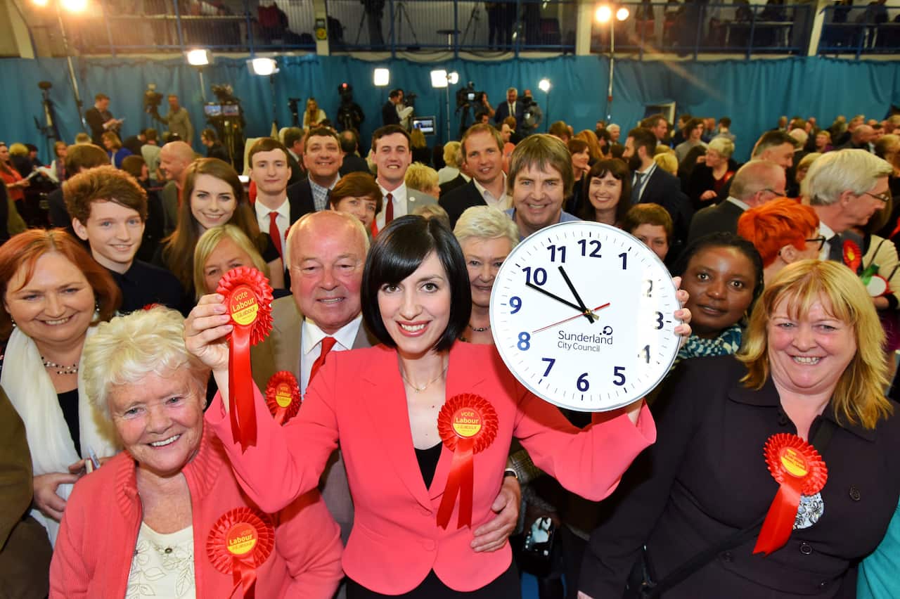 A woman wearing a peach coloured jacket holds a clock reading 10:50 aloft. 
