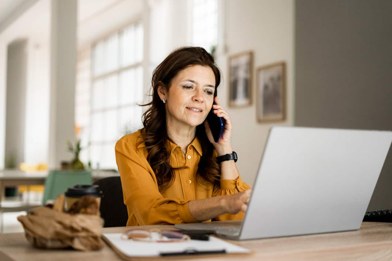 A smiling woman using a laptop while talking on her mobile phone at desk in home office.