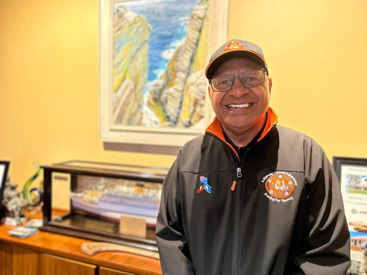 A man wearing a dark zip-up top and a cap and glasses standing and smiling inside 