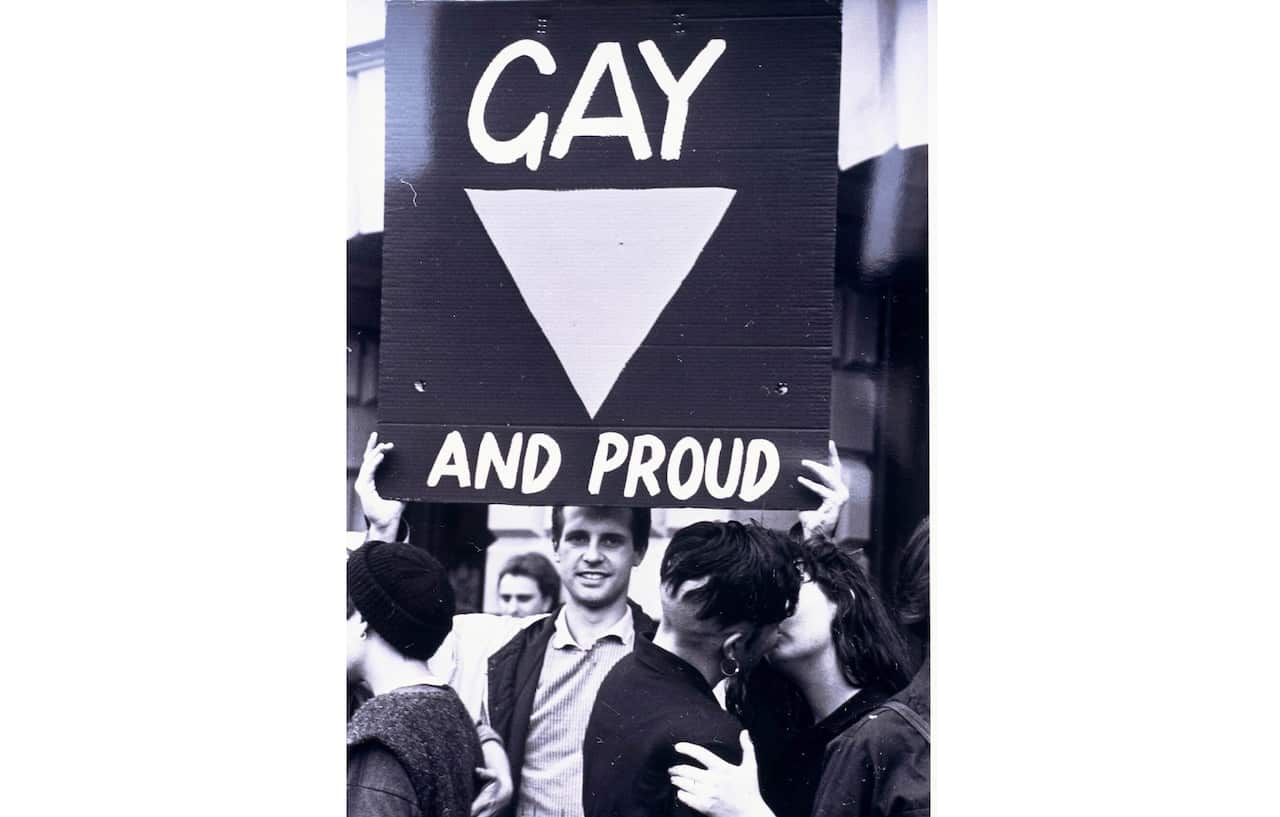A man holds a sign reading 'gay and proud' as two people kiss in front of him. 