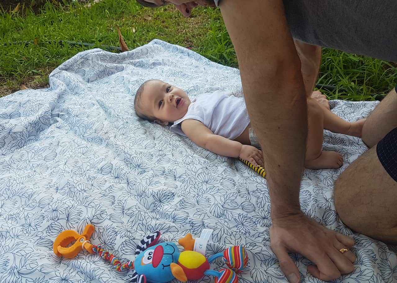 A young father leans over his happy baby, who lies on his back on a rug on the grass.