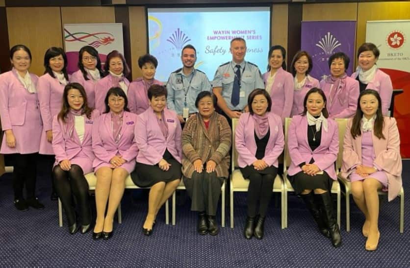 A row of women sitting in chairs inside. A row of women and two male police officers are standing behind them