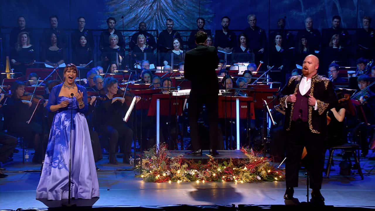 A man and a woman stand on stage, singing in front of an orchestra and choir. The ocnductor's stand is decorated with festive lights.