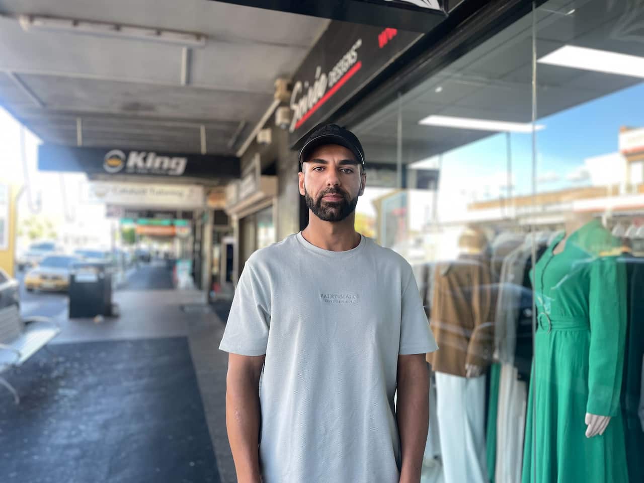 A young man wearing a white t-shirt and cap stands in front of shopfronts
