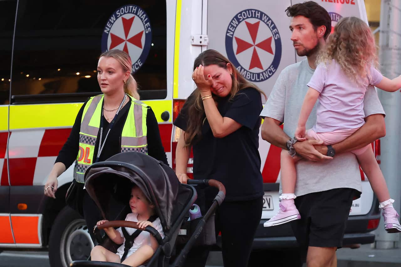 A woman rubs tears from her eyes as she pushes a pram, flanked by a police officer on one side and a man holding a small child in the other. Ambulance in the background.
