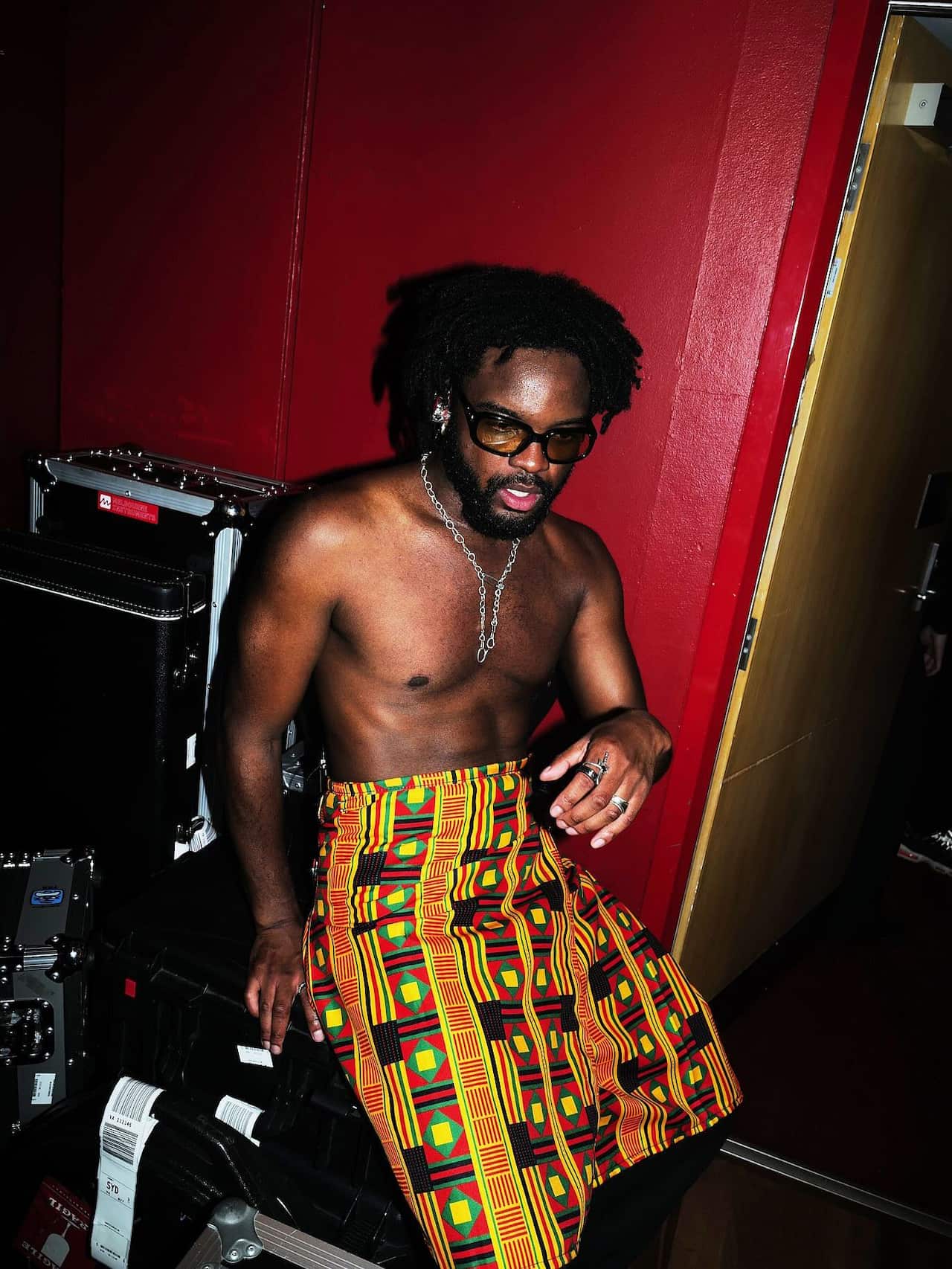 A shirtless man wearing a necklace and a yellow, green and red cloth wrap around his waist, is sitting on a case with musical equipment in the background