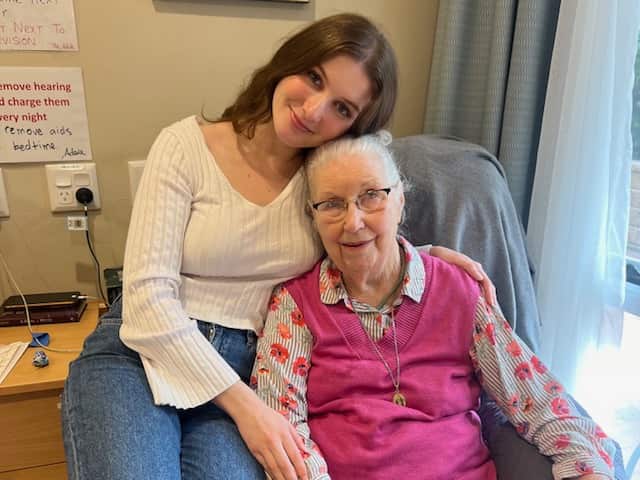 A young woman sitting on the seat of a chair that an older woman, her grandmother, is sitting on.