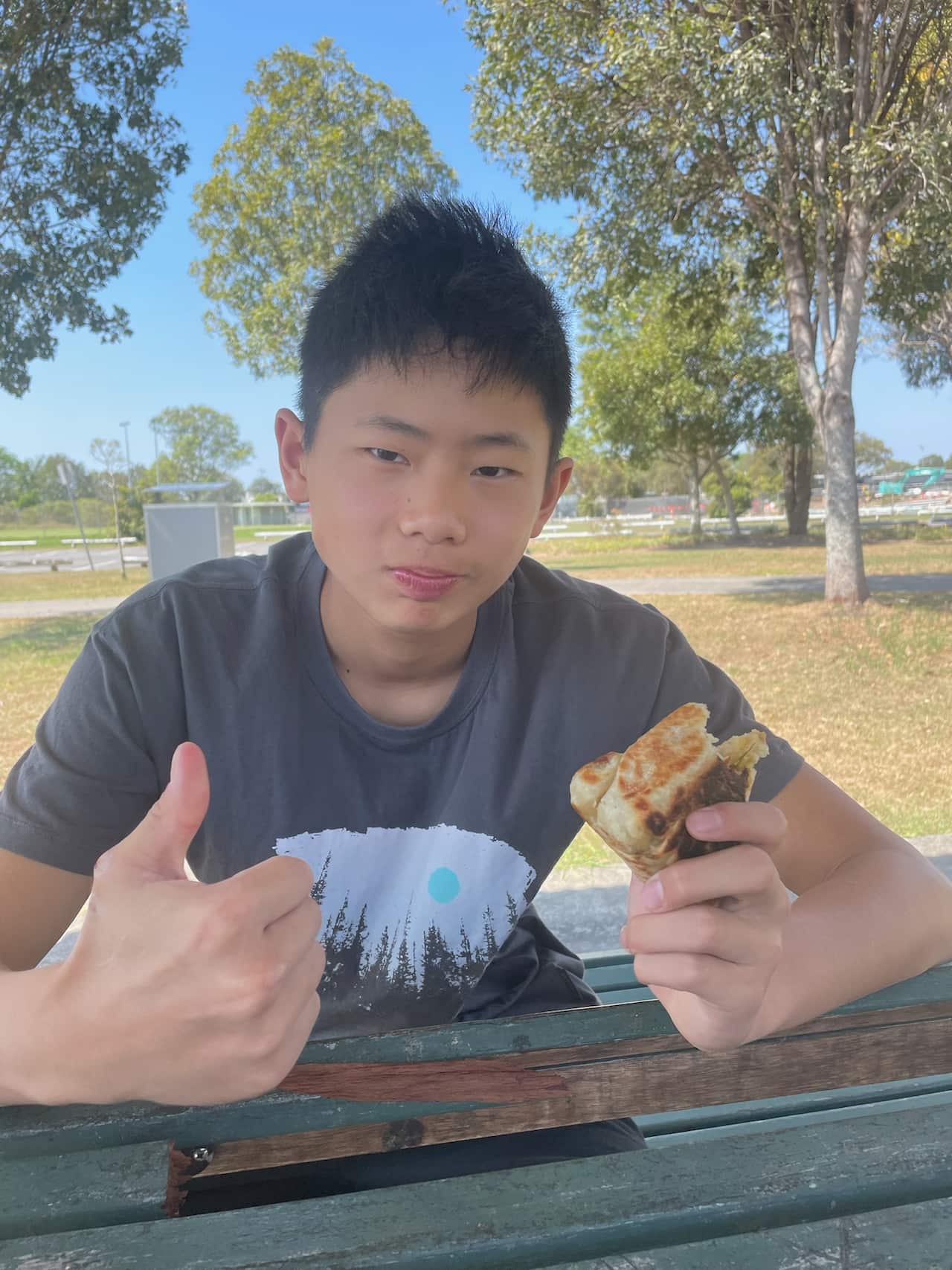 A young man sits at an outdoor park bench, giving a thumbs-up while holding a pastry. 