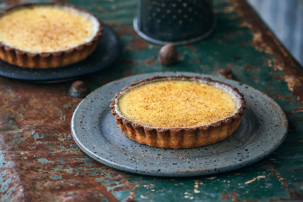 A round flat tart with a fluted pastry edge and golden centre sits on a flecked blue plate, with a second tart partially seen behind it.