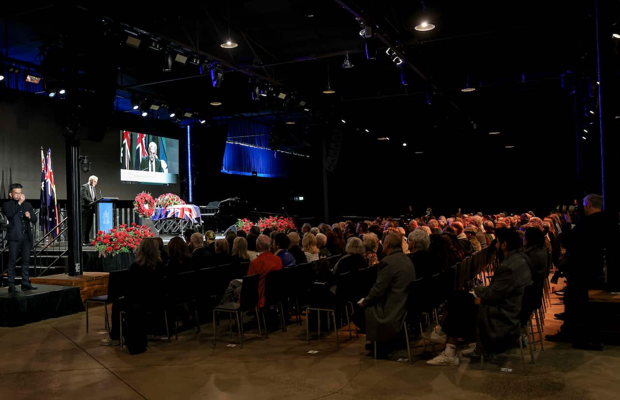A large crowd of people sitting in rows of seats, behind a stage with a casket on it.