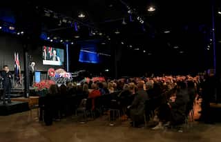 A large crowd of people sitting in rows of seats, behind a stage with a casket on it.