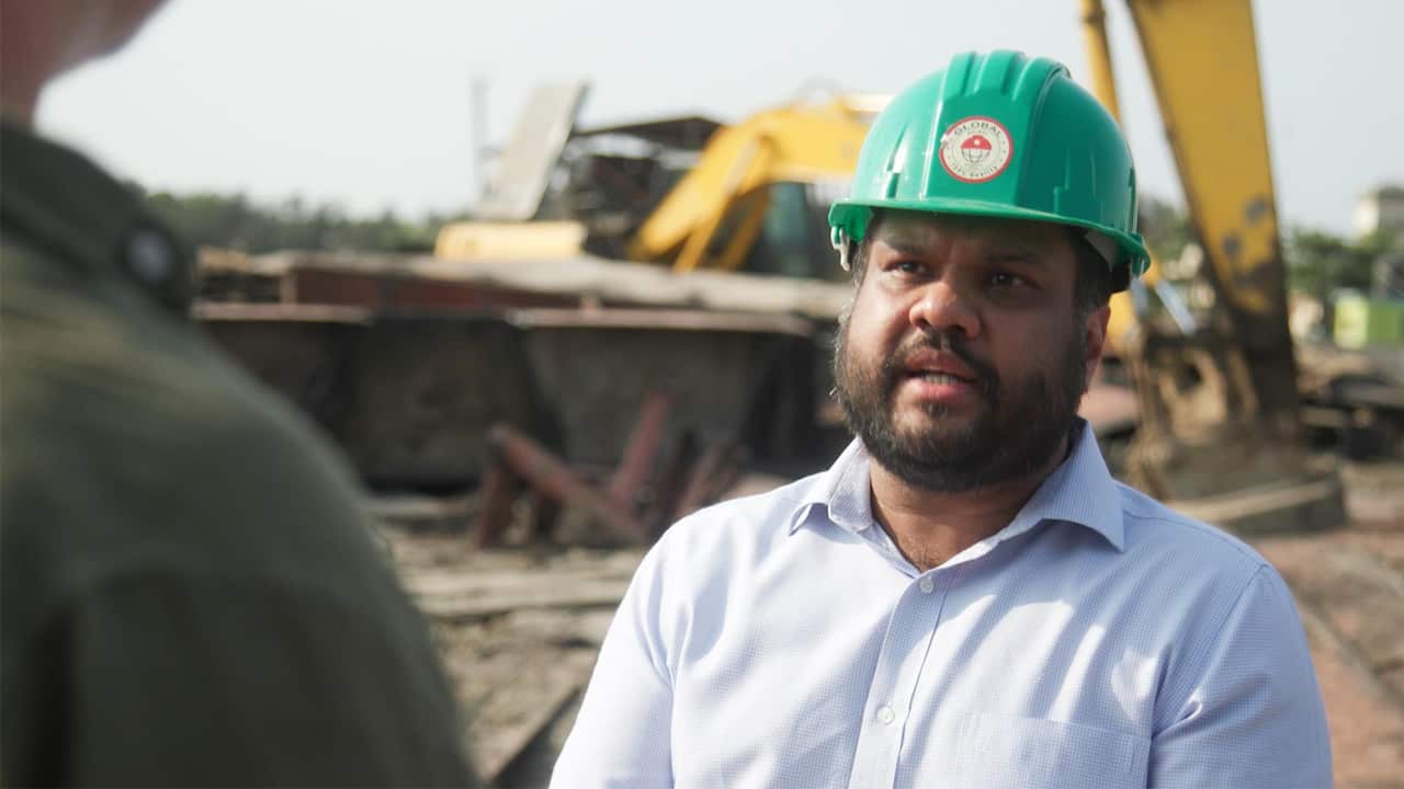 A South Asian man wearing a white shirt and a green safety helmet is talking to another person, who's only partially visible from behind