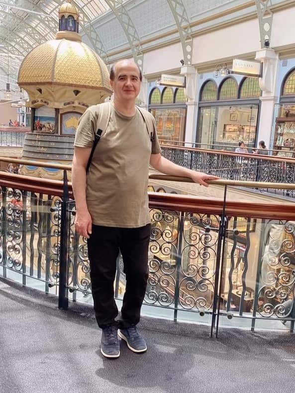 A balding man with dark hair and a light olive-green t-shirt stands on a second-story balcony of a large, ornate shopping arcade. He is looking at the camera and smiling faintly while his left hand rests on the decorative railing.