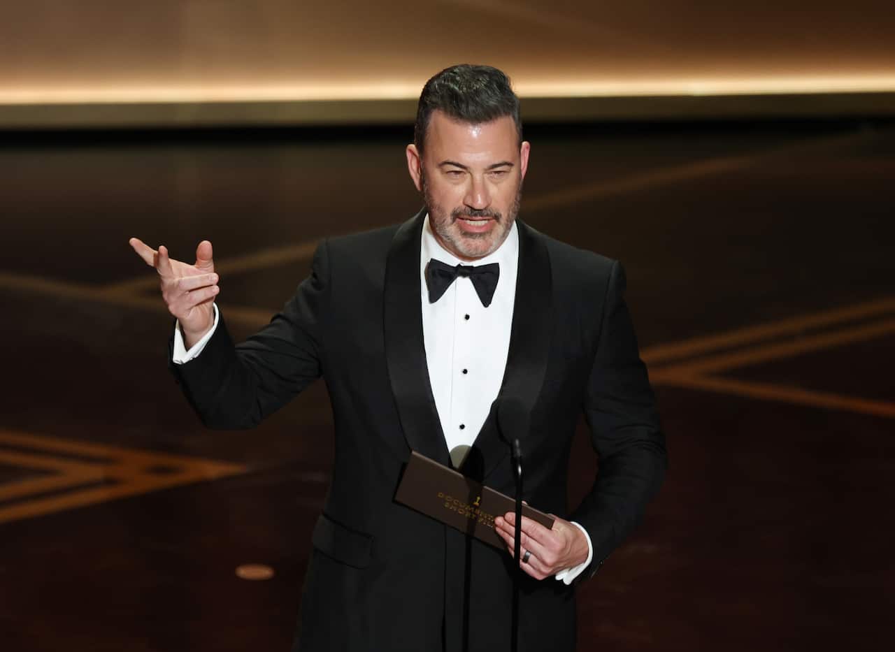 Jimmy Kimmel, standing on stage, holding an envelope and speaking into a microphone.