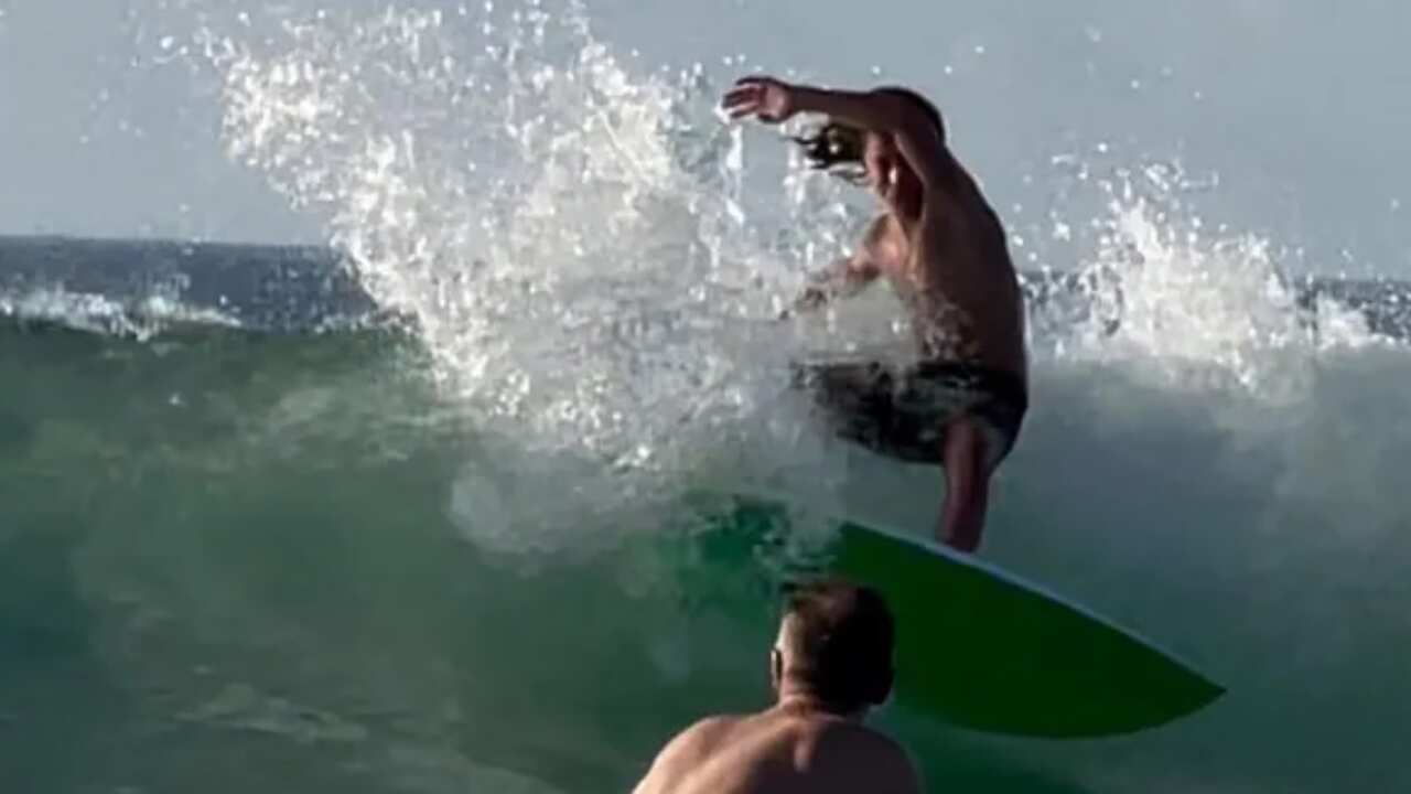 Khai Cowley surfs on the crest of a wave while another surfer in the foreground watches on.
