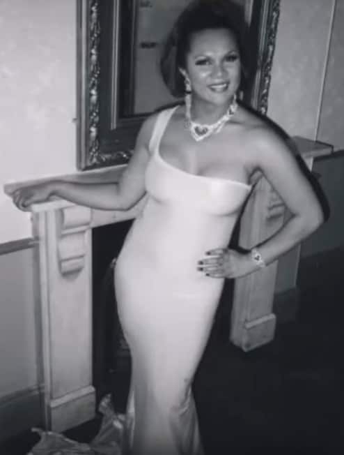 A blak and white photo of a woman smiling, wearing a black and white dress and leaning away from a wall with a fireplace.