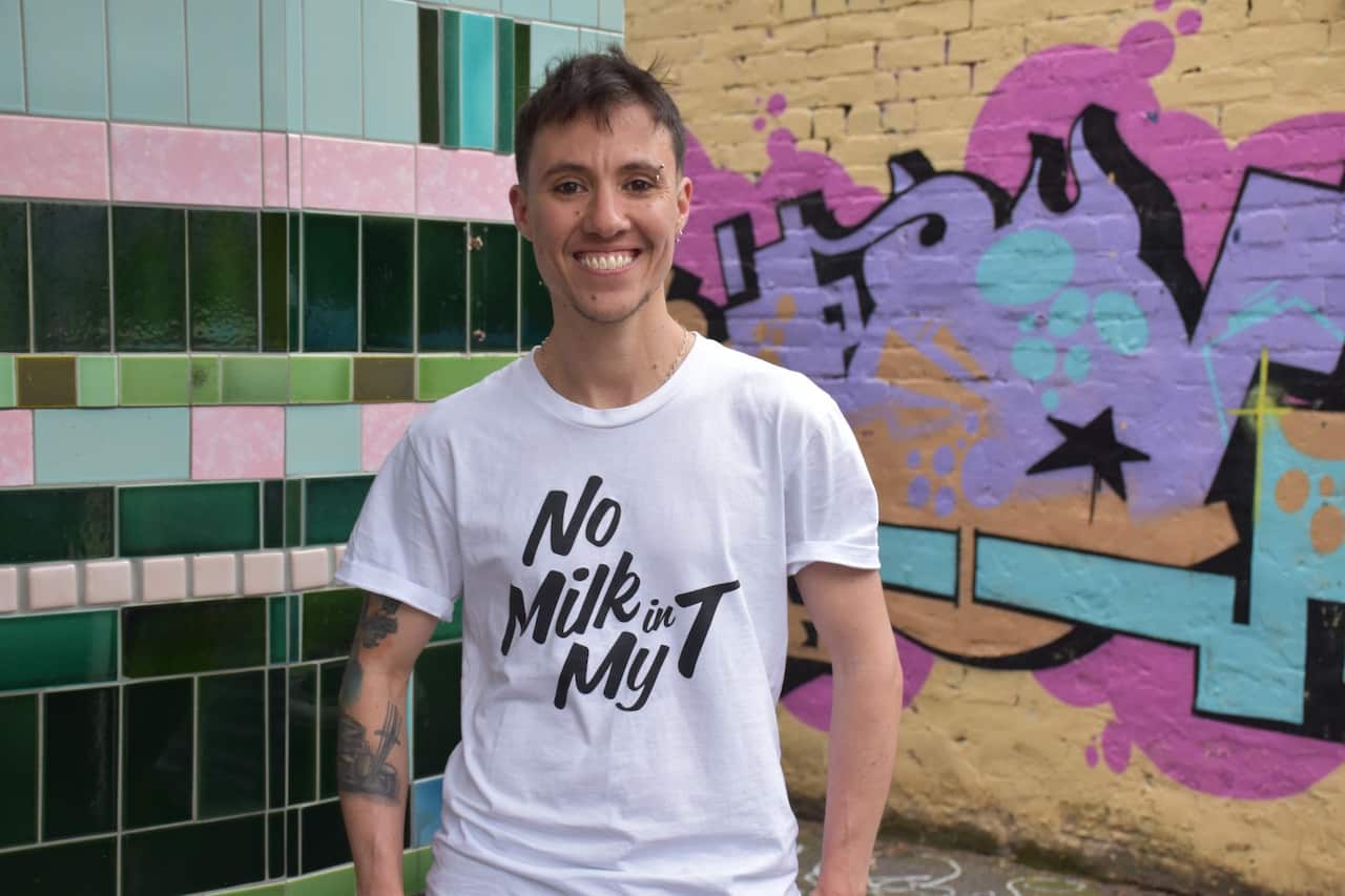 A person with short brown hair and an eyebrow piercing poses for a photo wearing a white T-shirt with the words No Milk in My T printed on the front in a stylised italic black font. 