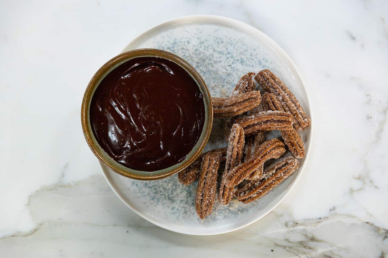 A bowl of glossy chocolate sauce sits on a wide, speckled plate. Beside the bowl, on the plate, sit a pile of ridged long, brown pastry strips.