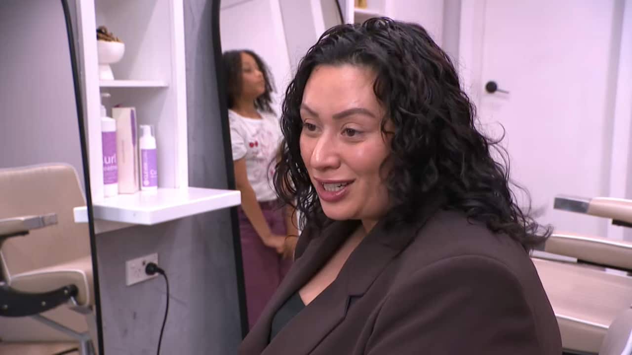 A woman in a brown blazer with curly hair is talking as she sits inside a salon. There's a young girl with curly hair standing behind her.
