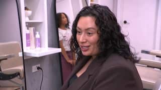 A woman in a brown blazer with curly hair is talking as she sits inside a salon. There's a young girl with curly hair standing behind her.
