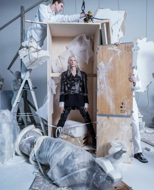 A woman in a studded leather jacket stands in a wooden crate. In front of her is a statue wrapped in white material