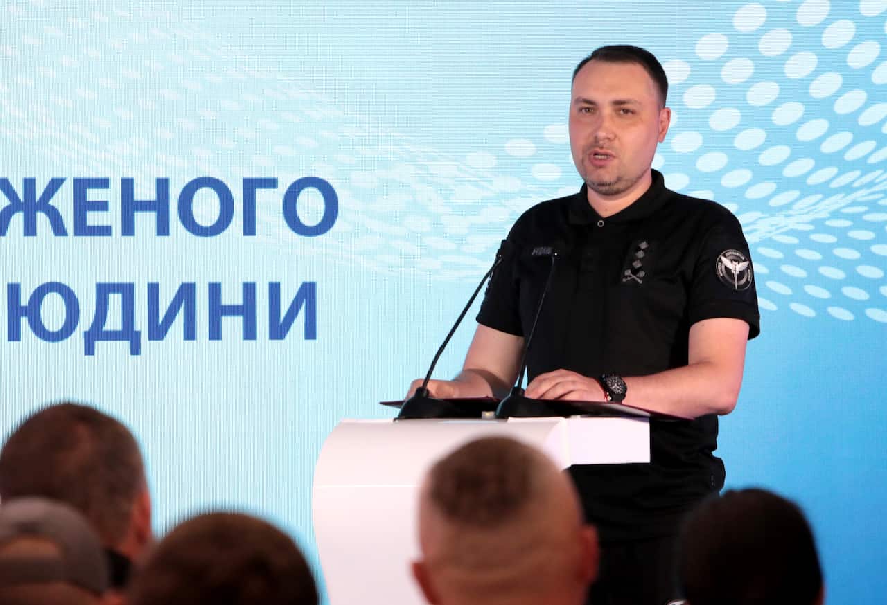 A man in a black polo shirt speaks at a podium with microphones in front of a blue backdrop with Ukrainian text.