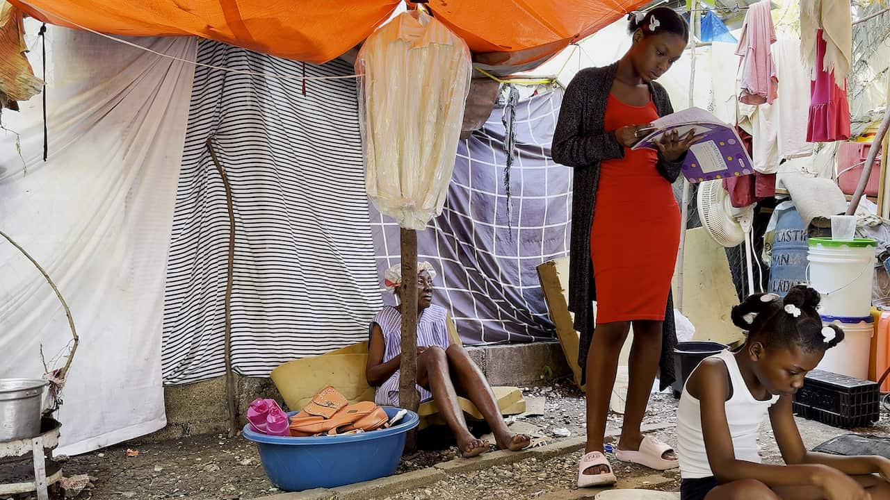 Two girls, one of them a teenager holding a school book, and an elderly woman sit inside a makeshift tent.