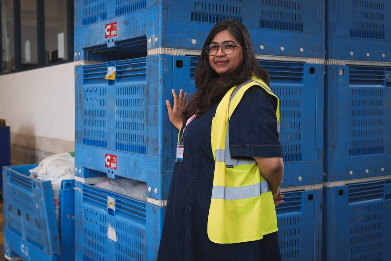 A woman with a safety vest is posing in front of a number of blue crates filled with clothing bags