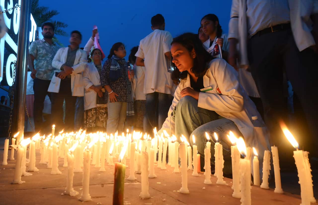 A large amount of lit candles on a table during a vigil. One person is kneeling down near them, others are standing up further away from them.