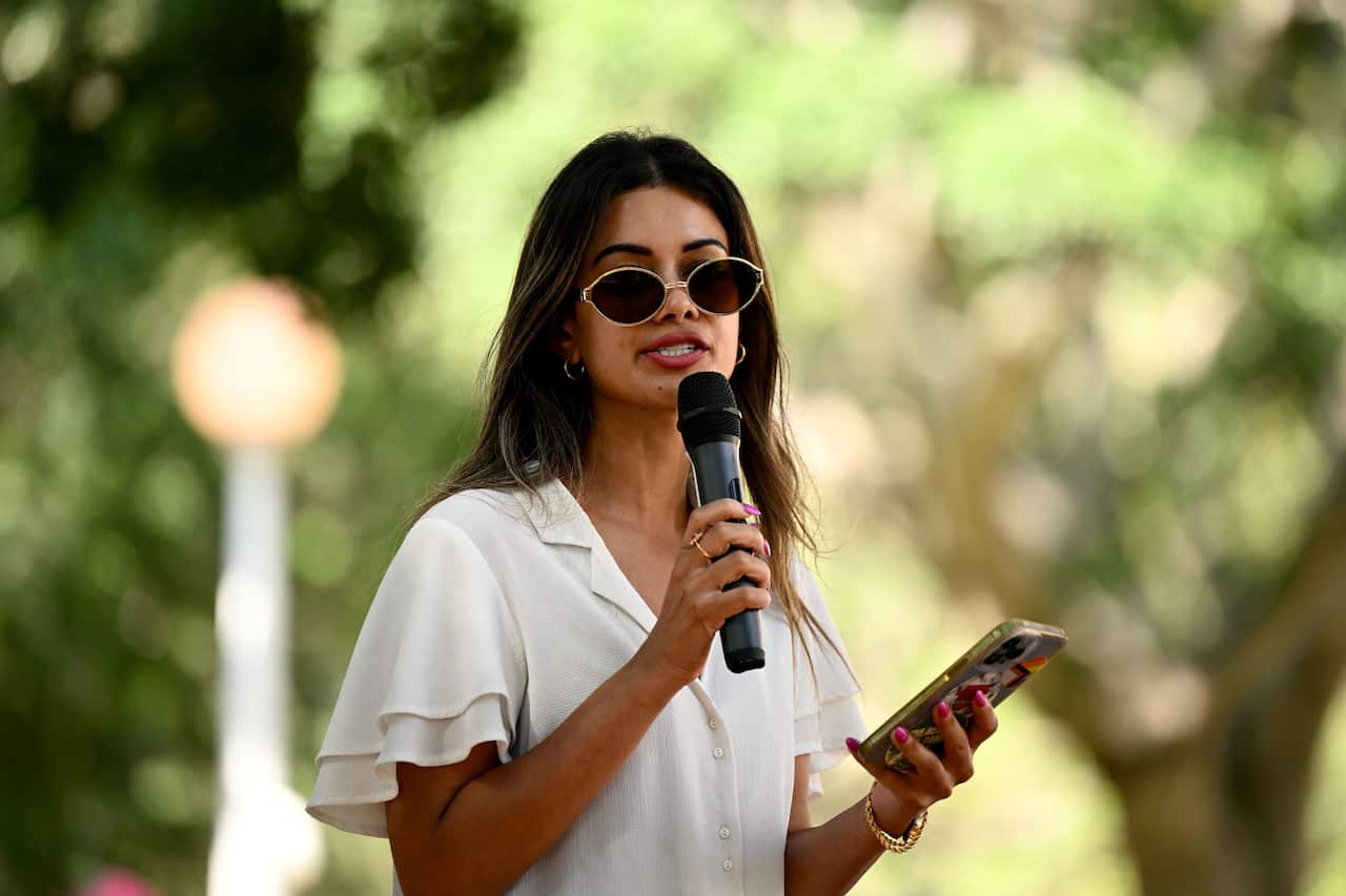 A woman wearing a white top and sunglasses, speaking into a microphone she's holding in one hand. She's holding a mobile phone in her other hand.