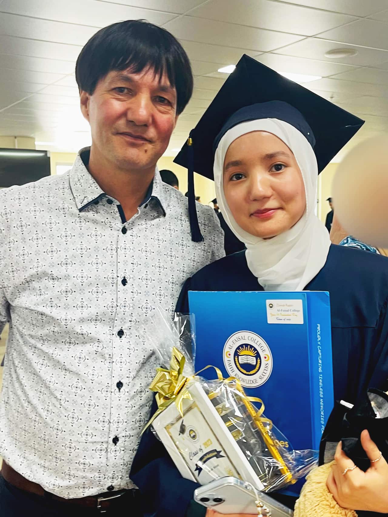 A father wearing a white collared shirt poses with his daughter in a university graduation gown and mortar board