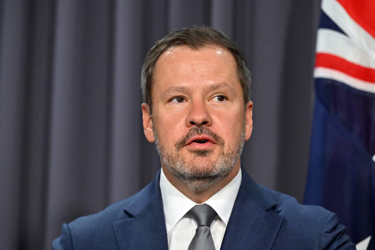 A man speaking in front of a blue curtain and Australian flag.