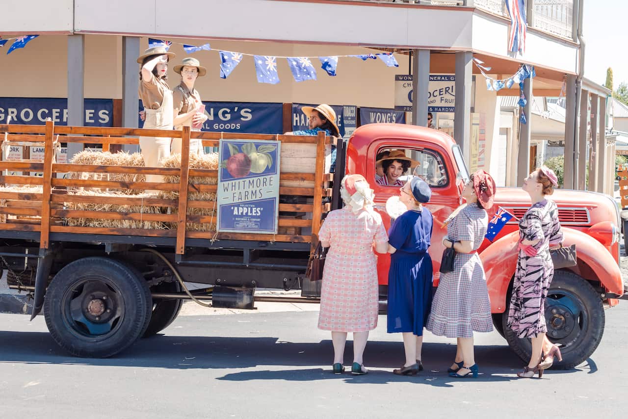 A truck from an Apple farm drives through a town, four women look up at a lady saluting from the trailer of the truck