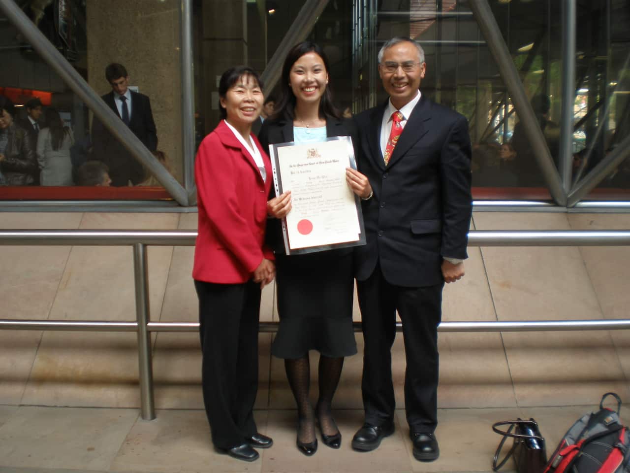 A young Asian woman holds a university certificate while posing with her smiling parents 