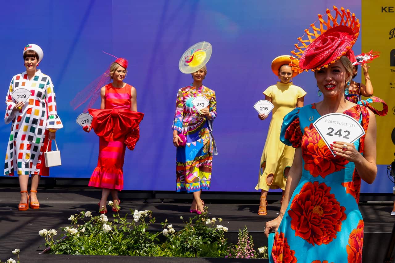 Five women on stage dressed up in dresses and fascinators. They are competing in a fashion contest.
