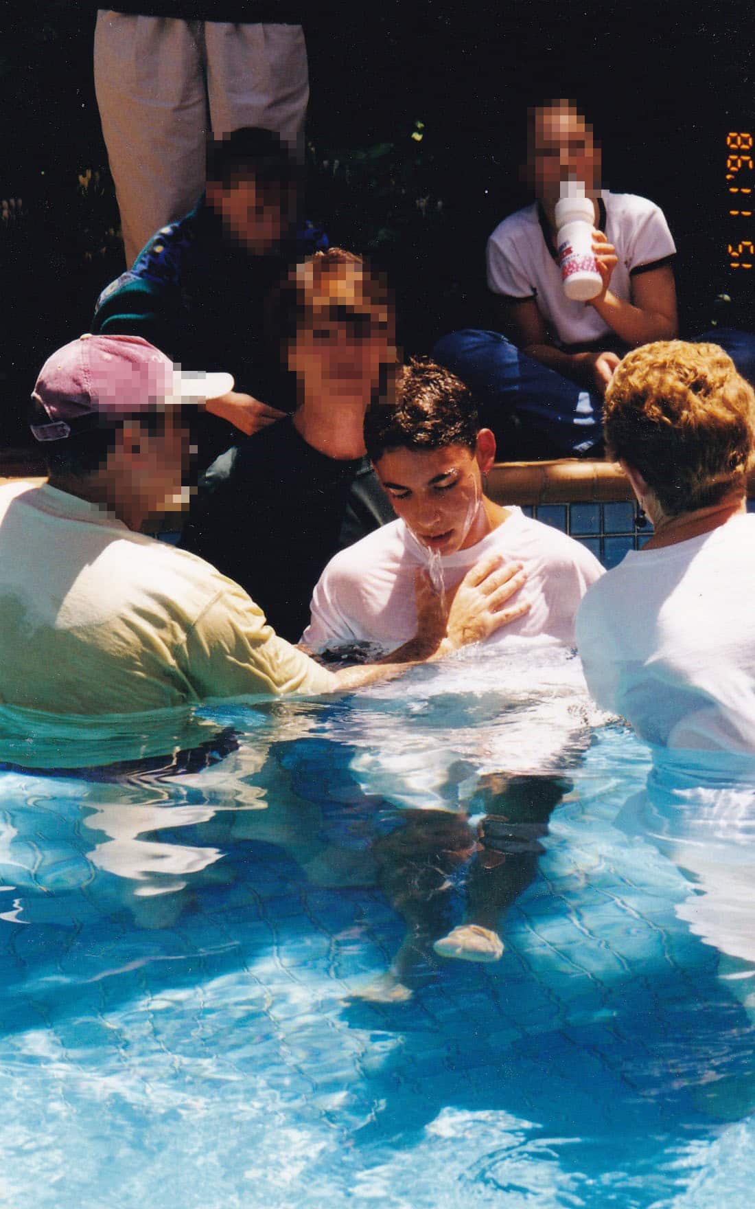 A man in a white -shirt about to be submerged in a swimming pool. There are people around him in and out of the pool.