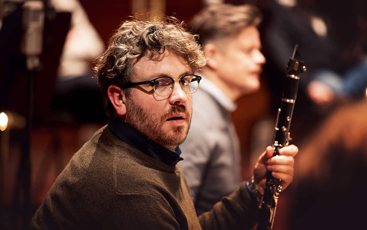 Man with curly hair and glasses in brown jersey holds clarinet, turning to look sideways.