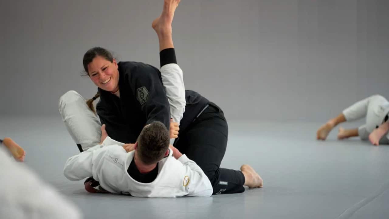 a woman with long brown hair in a braid and in a black uniform trains in jiu-jitsu with a man in white on the ground